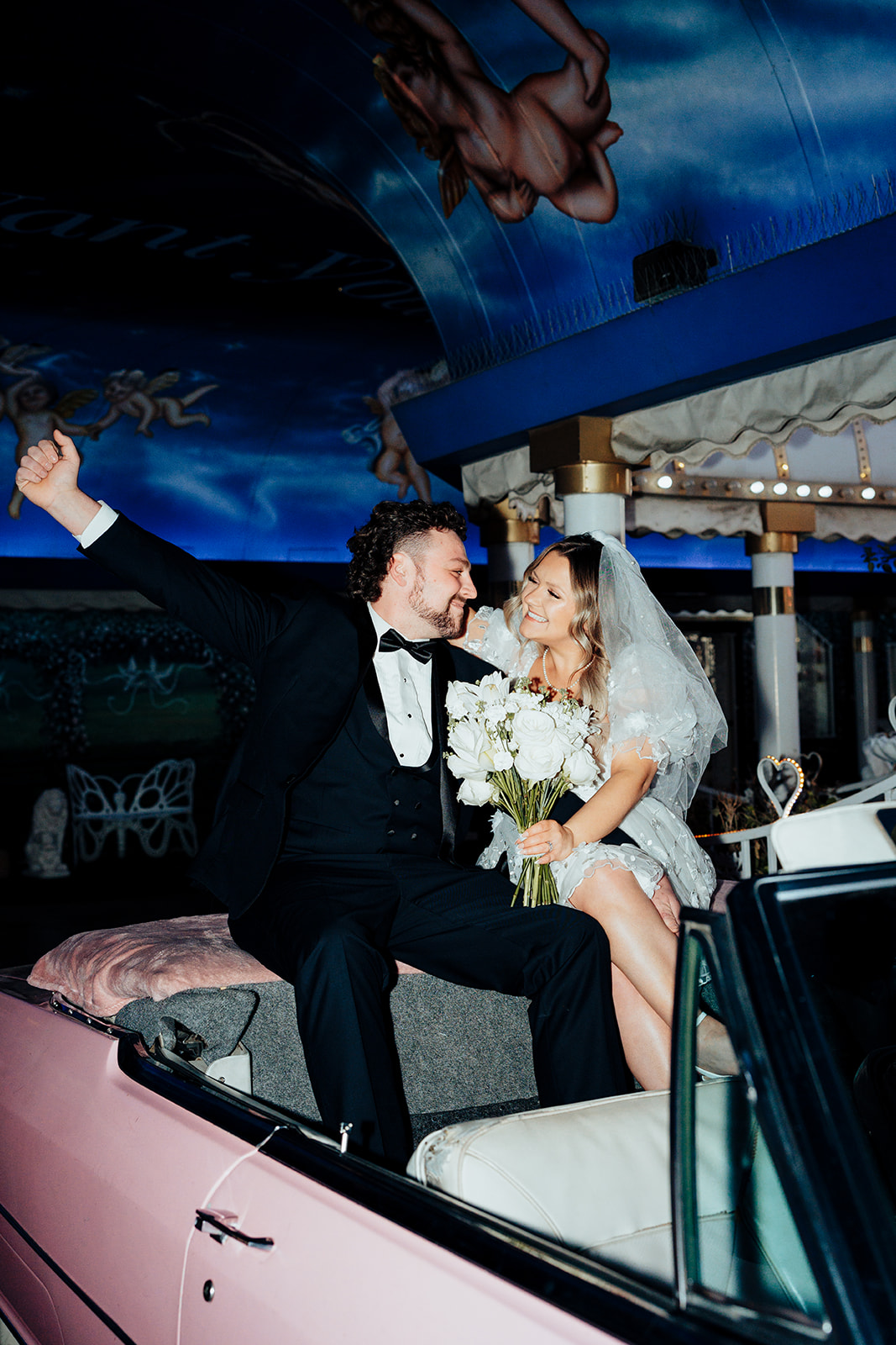 Bride and groom in a pink car celebrating their elopement at Little White Wedding Chapel, with floral bouquet and blue ceiling decor.