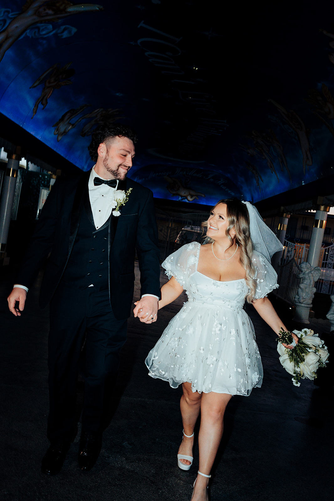 Bride and groom walking hand in hand inside Little White Wedding Chapel after elopement, with blue ceiling and angelic decor in the background.