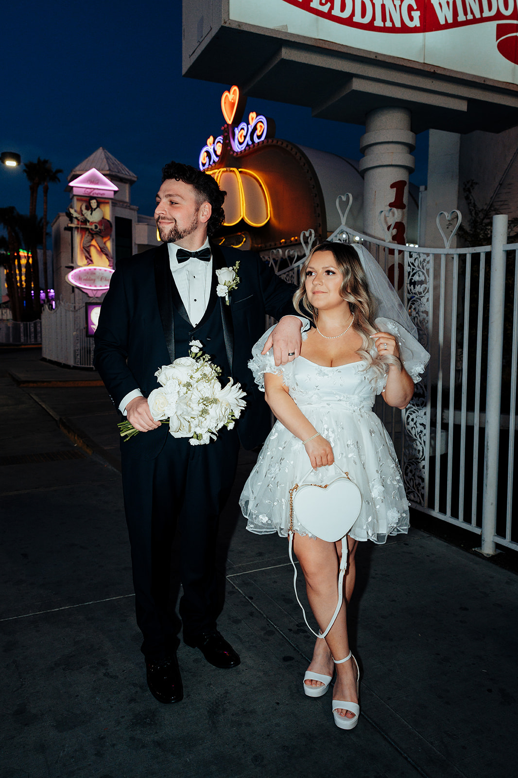 Bride and groom posing in front of Little White Wedding Chapel’s Wedding Window sign at night, with neon lights and a floral bouquet.