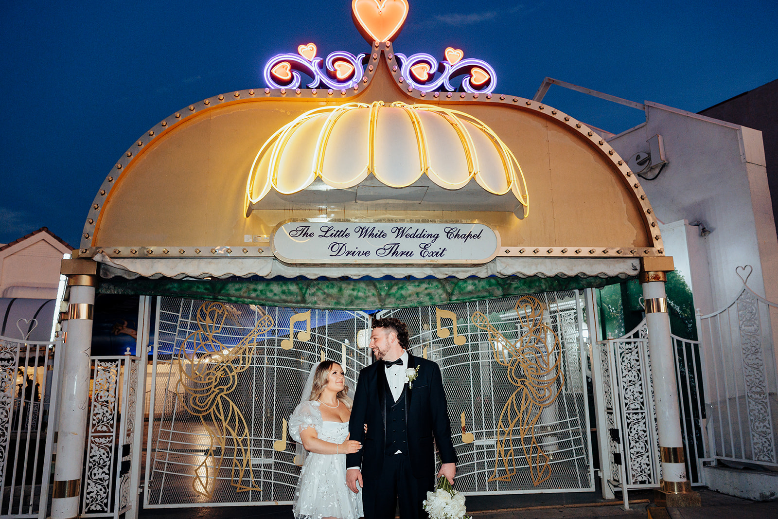 Bride and groom posing in front of Little White Wedding Chapel Drive-Thru Exit sign after elopement, with neon lights and floral bouquet at night.