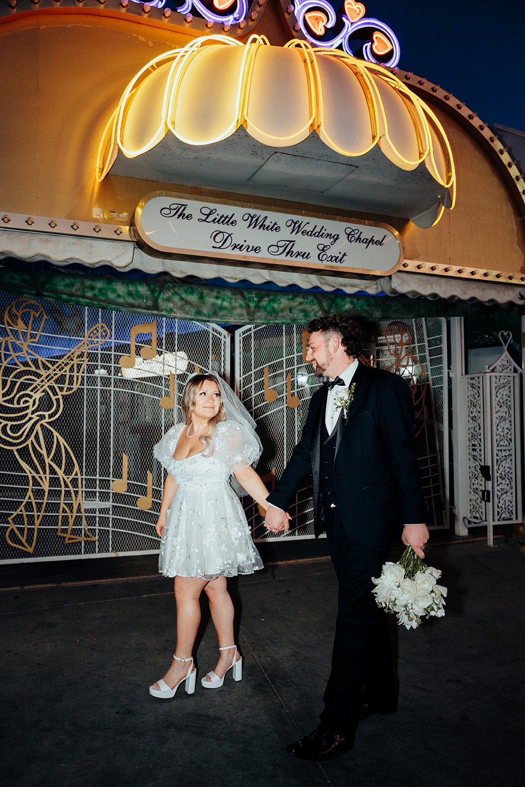 Bride and groom walking hand in hand at Little White Wedding Chapel Drive-Thru Exit after elopement, with neon lights and floral bouquet at night.