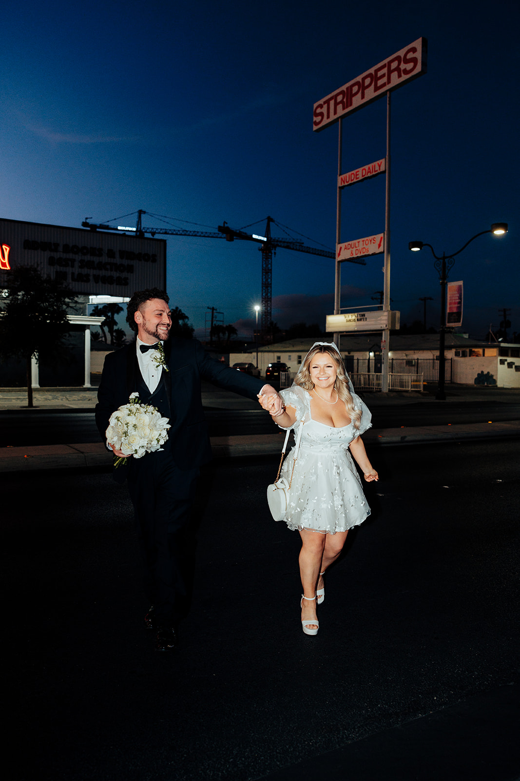 Bride and groom walking hand in hand after Little White Wedding Chapel elopement on Las Vegas street, with 'Stripper' sign and construction cranes in the background.