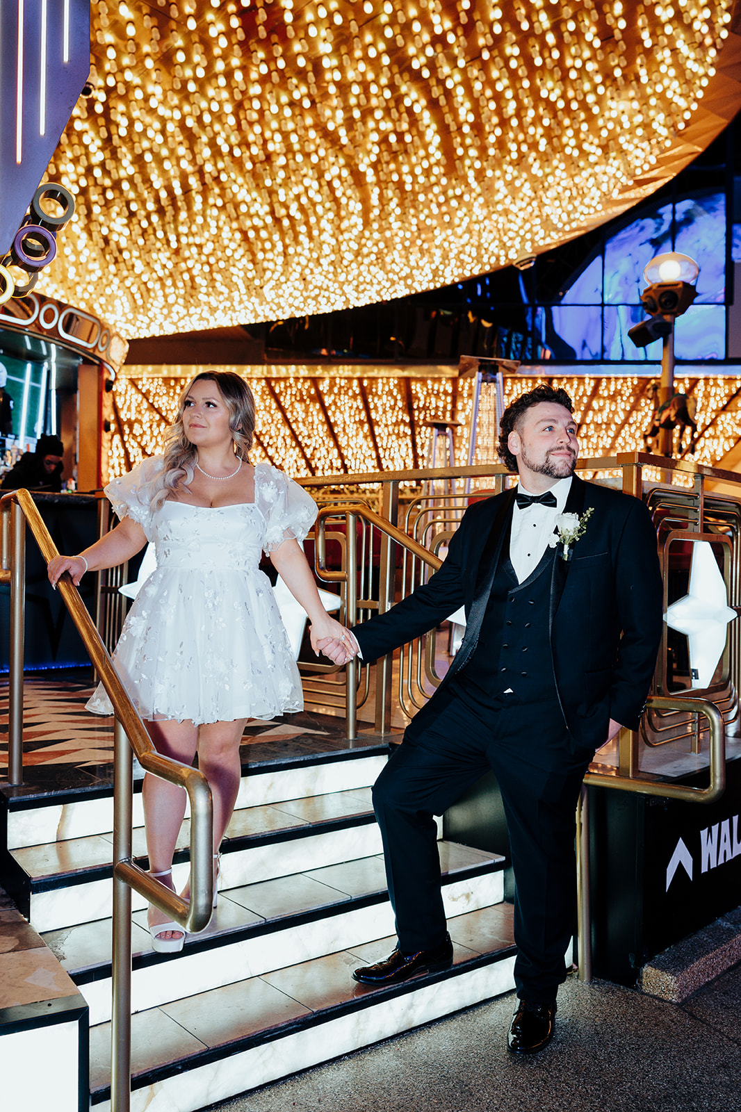 Bride and groom walking down staircase on Las Vegas Strip after Little White Wedding Chapel elopement, with bright ceiling lights in the background.