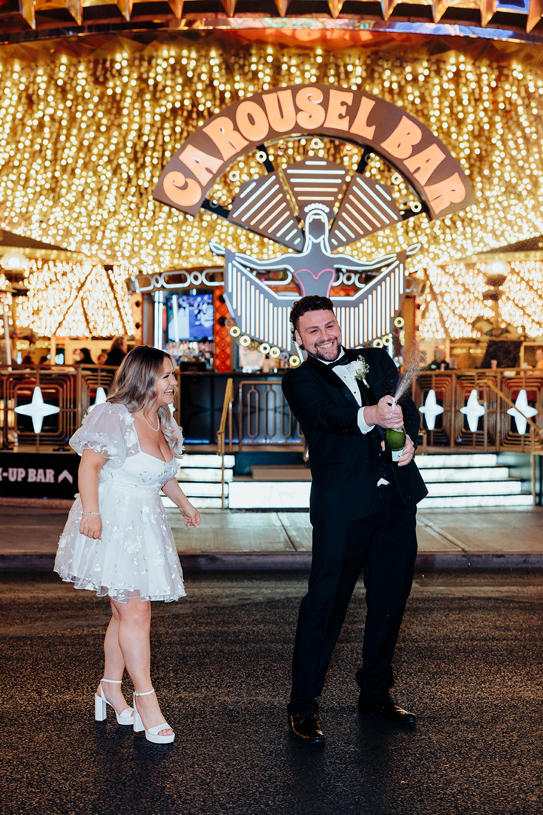 Groom popping champagne on Las Vegas Strip after Little White Wedding Chapel elopement, with bride smiling under Carousel Bar sign and bright lights.