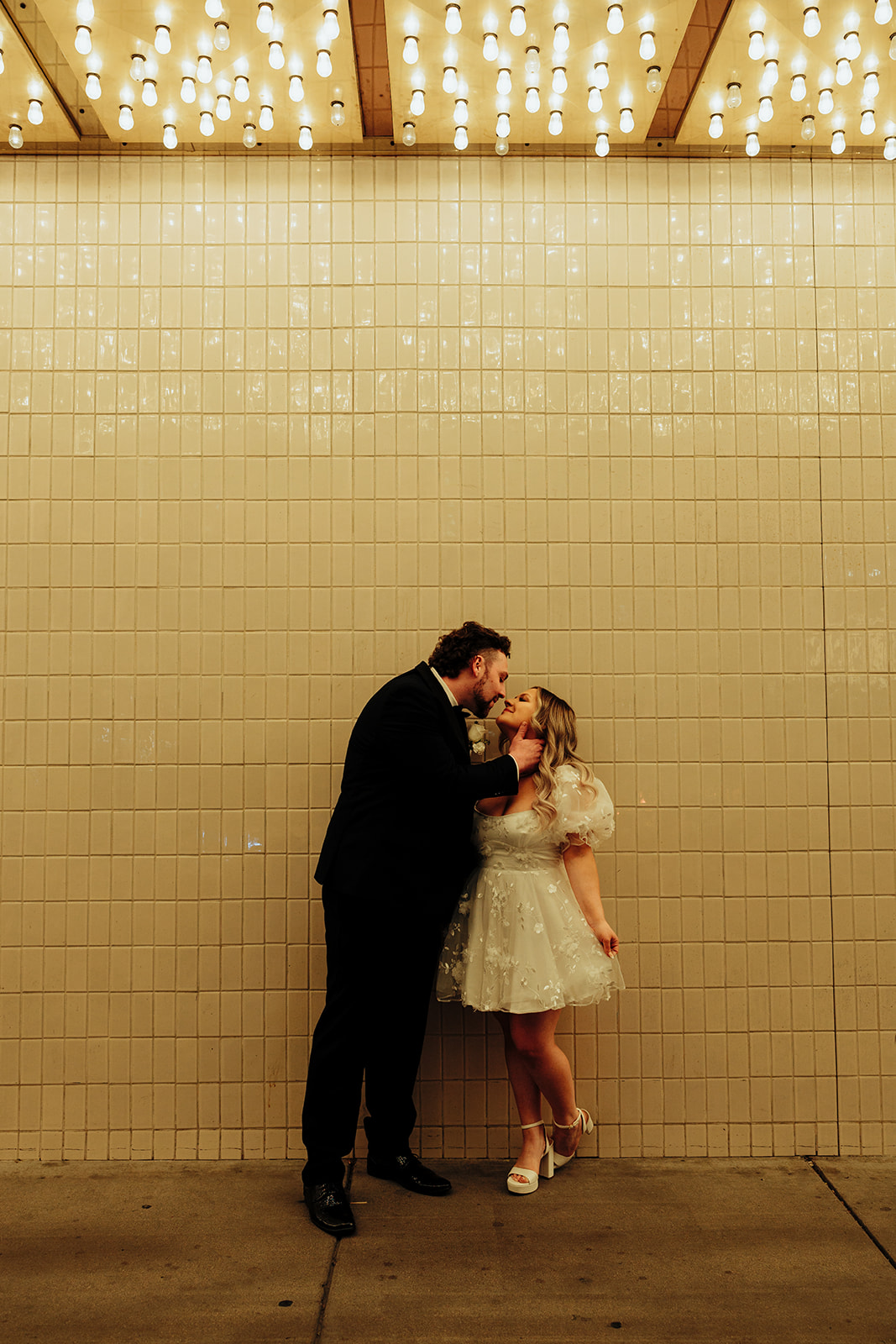 Bride and groom kissing under twinkling lights on Las Vegas Strip after Little White Wedding Chapel elopement, with tiled wall in the background.