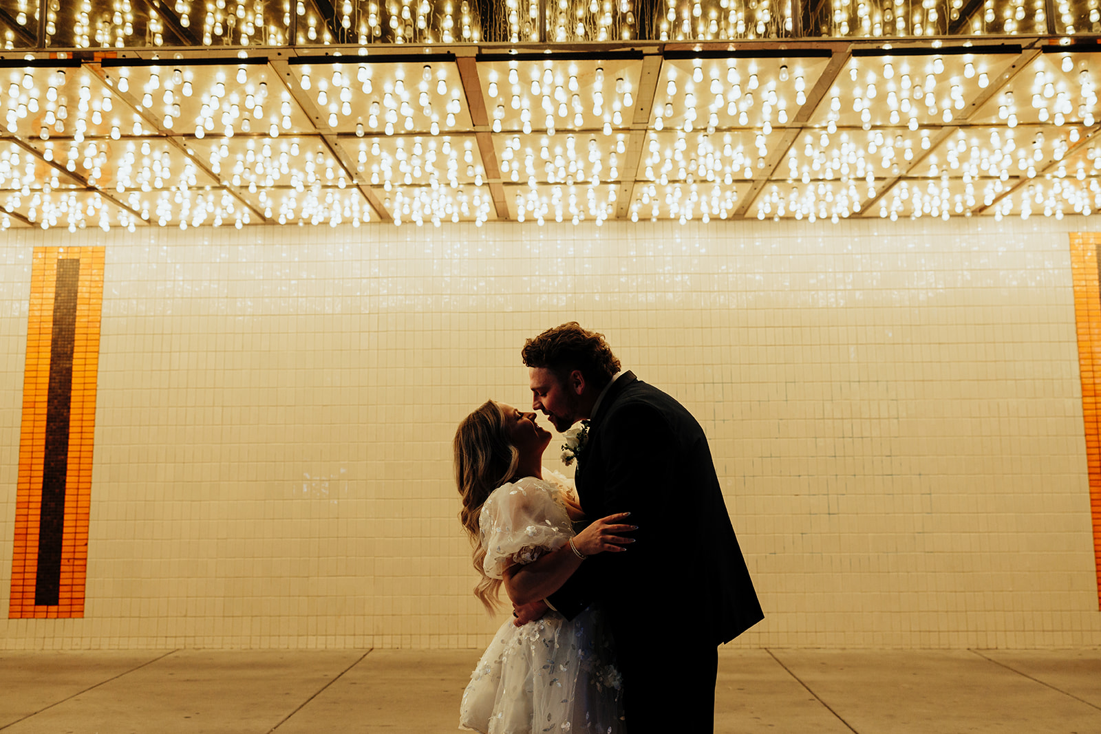 Bride and groom sharing a kiss under twinkling lights on Las Vegas Strip after Little White Wedding Chapel elopement, with tiled wall in the background.