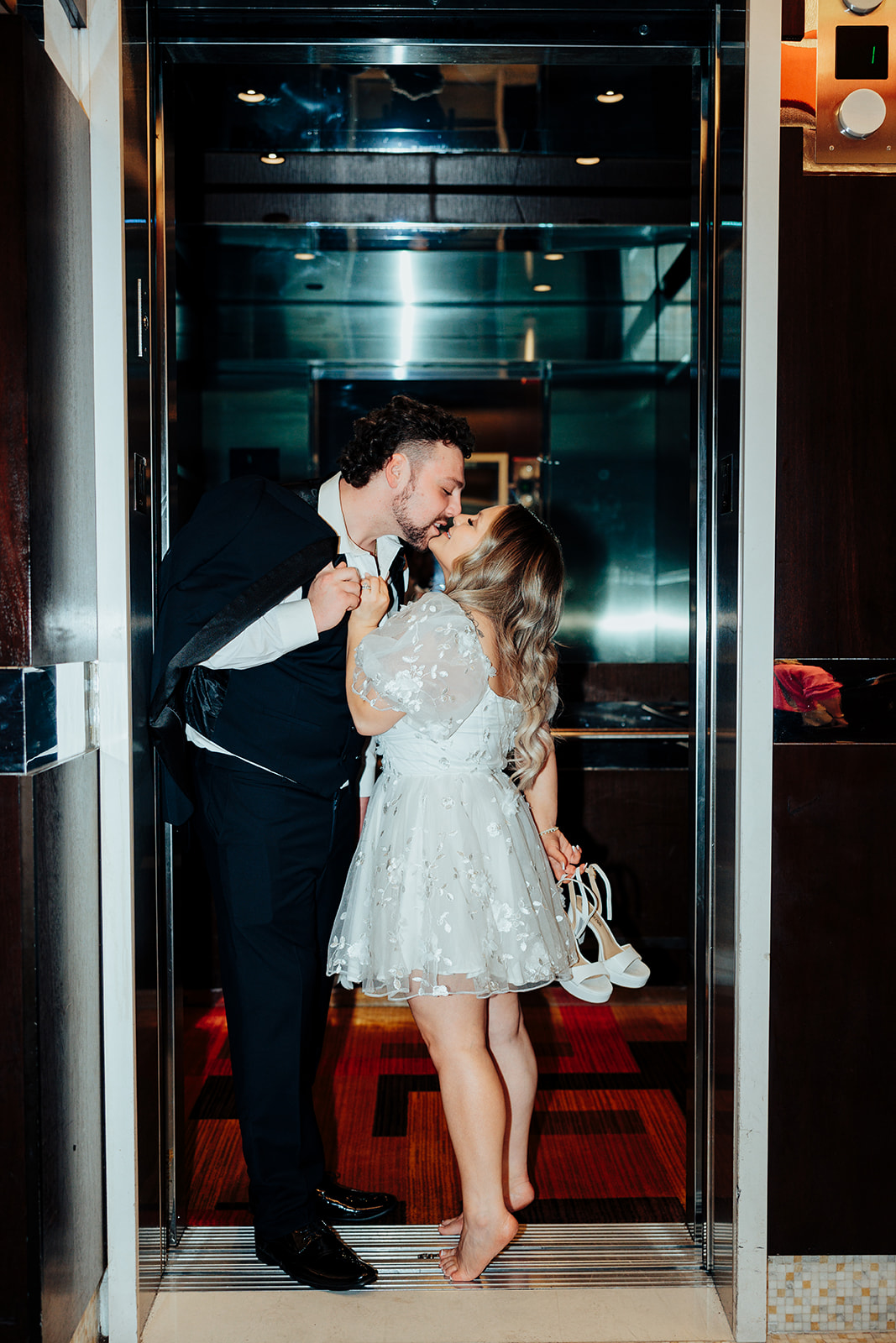 Bride and groom kissing in elevator at Golden Nugget after Little White Wedding Chapel elopement, with bride holding white heels.