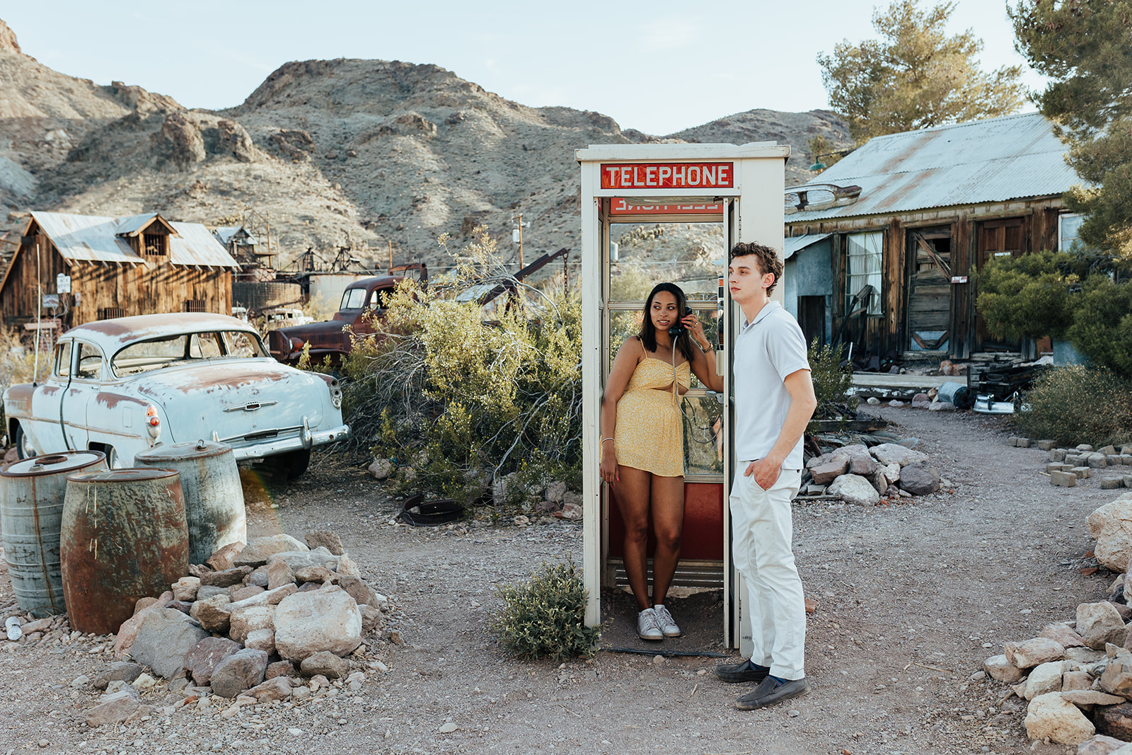 A couple posing by a vintage telephone booth at Nelson's Ghost Town, Nevada, with rusted cars, desert vegetation, and mountains in the background.