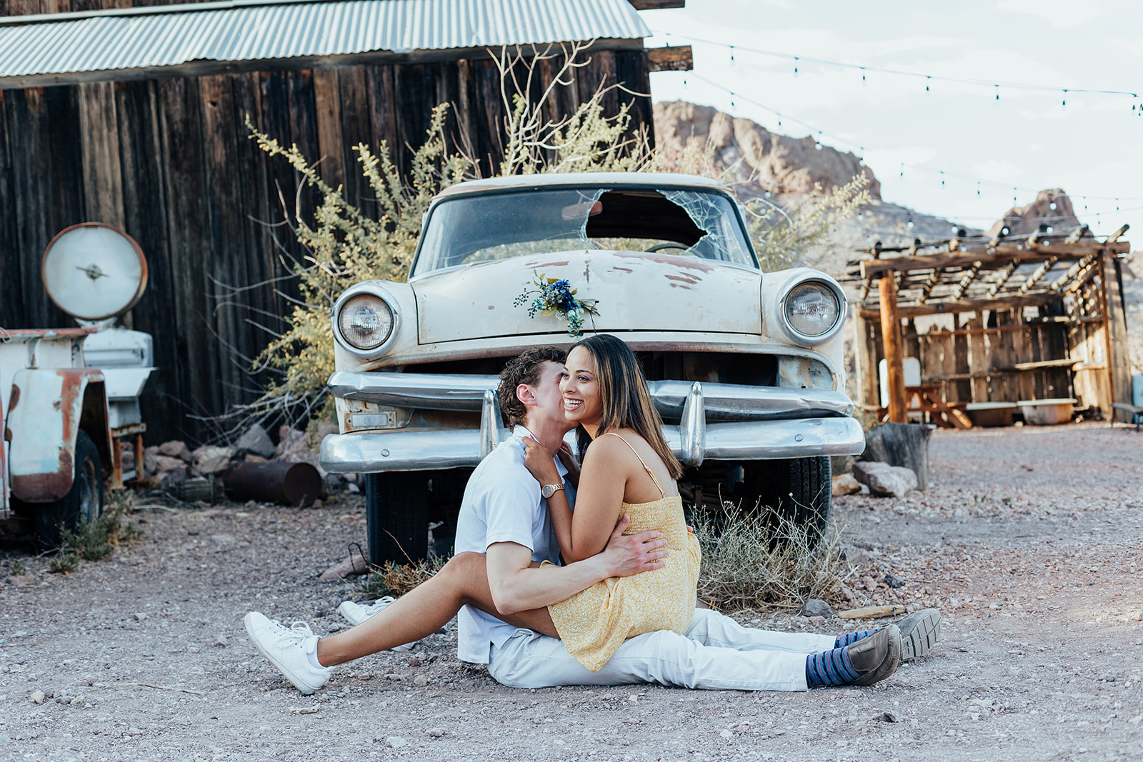 A couple embraces in front of a vintage car at Nelson's Ghost Town, surrounded by rustic desert scenery and string lights, capturing a joyful wedding proposal moment.