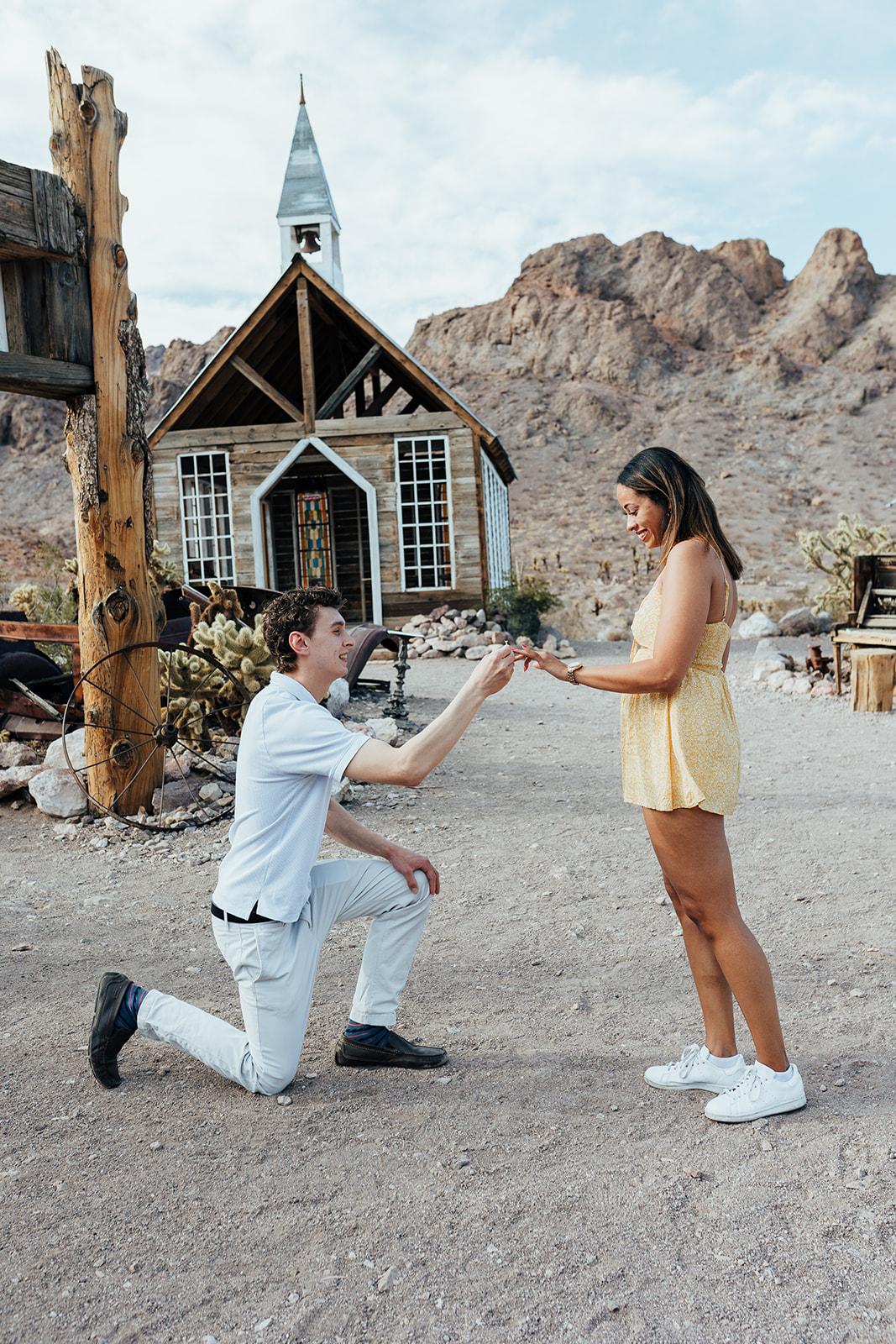 A person proposing on one knee in front of a small wooden chapel at Nelson's Ghost Town, Nevada, with a desert landscape and mountains in the background.
