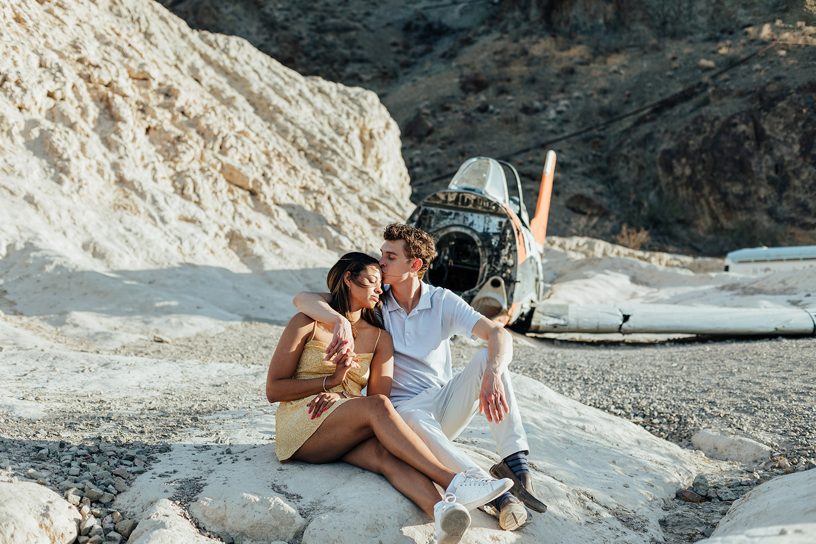 A couple shares a gentle kiss on a rock at Nelson's Ghost Town, with a wrecked plane in the background, capturing a romantic wedding proposal in a rugged desert landscape.