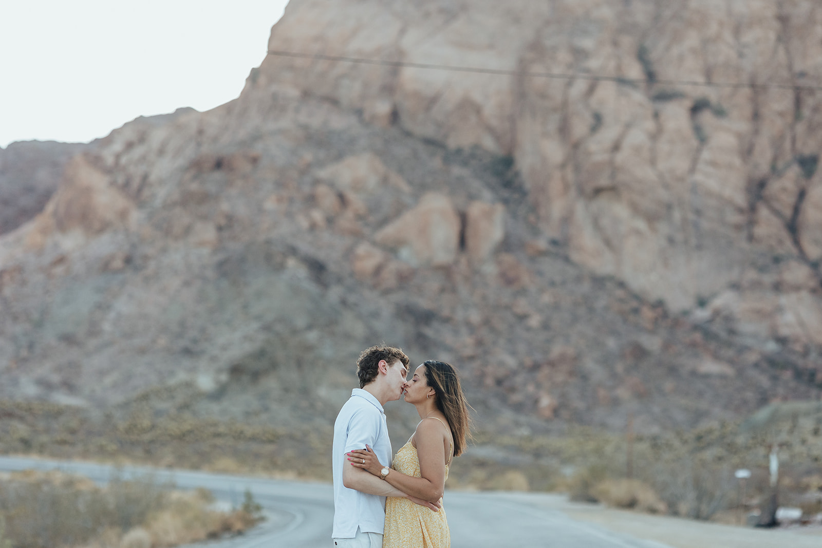 A couple shares a loving kiss on a deserted road at Nelson's Ghost Town, with dramatic desert cliffs in the background, capturing a romantic wedding proposal moment.