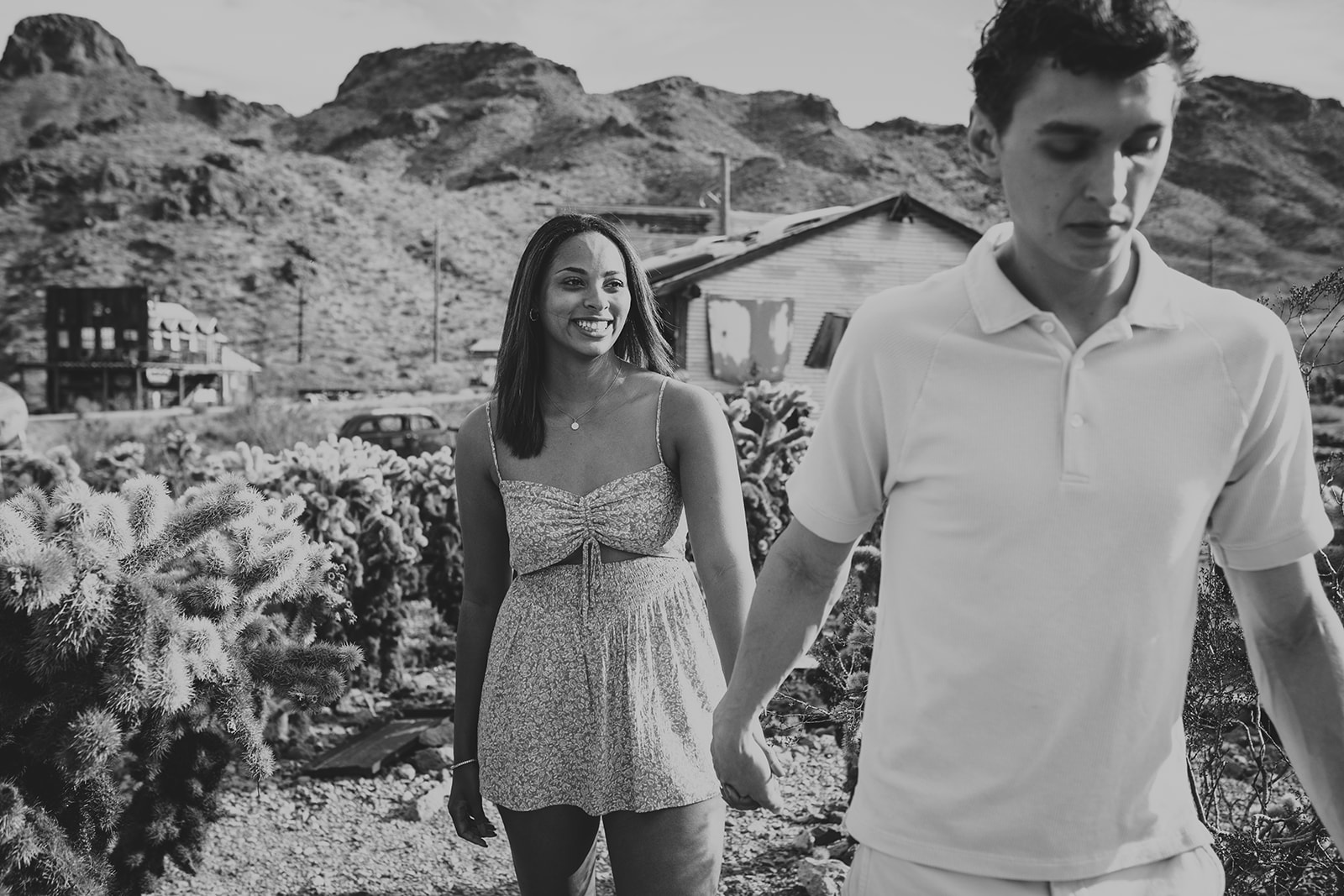 A couple holding hands in a black-and-white photo at Nelson's Ghost Town, Nevada, with cacti, vintage cars, and desert mountains in the background.
