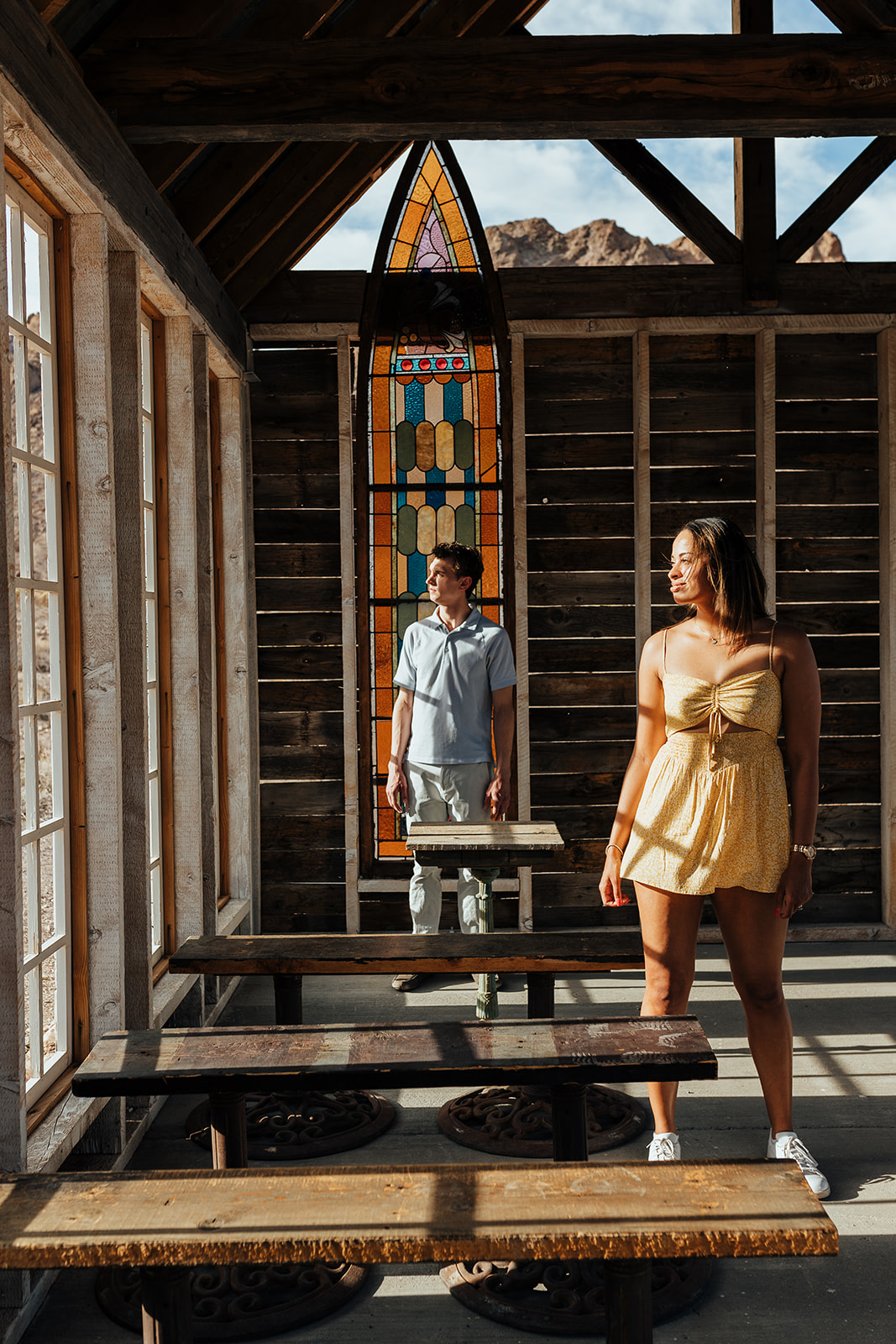 A couple posing inside a rustic chapel at Nelson's Ghost Town, Nevada, with a colorful stained glass window and desert mountains visible in the background.