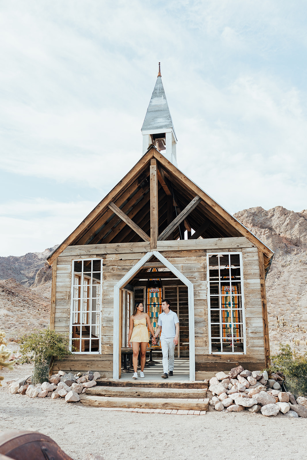 A couple standing at the entrance of a rustic wooden chapel at Nelson's Ghost Town, Nevada, with desert mountains and rocks in the background.