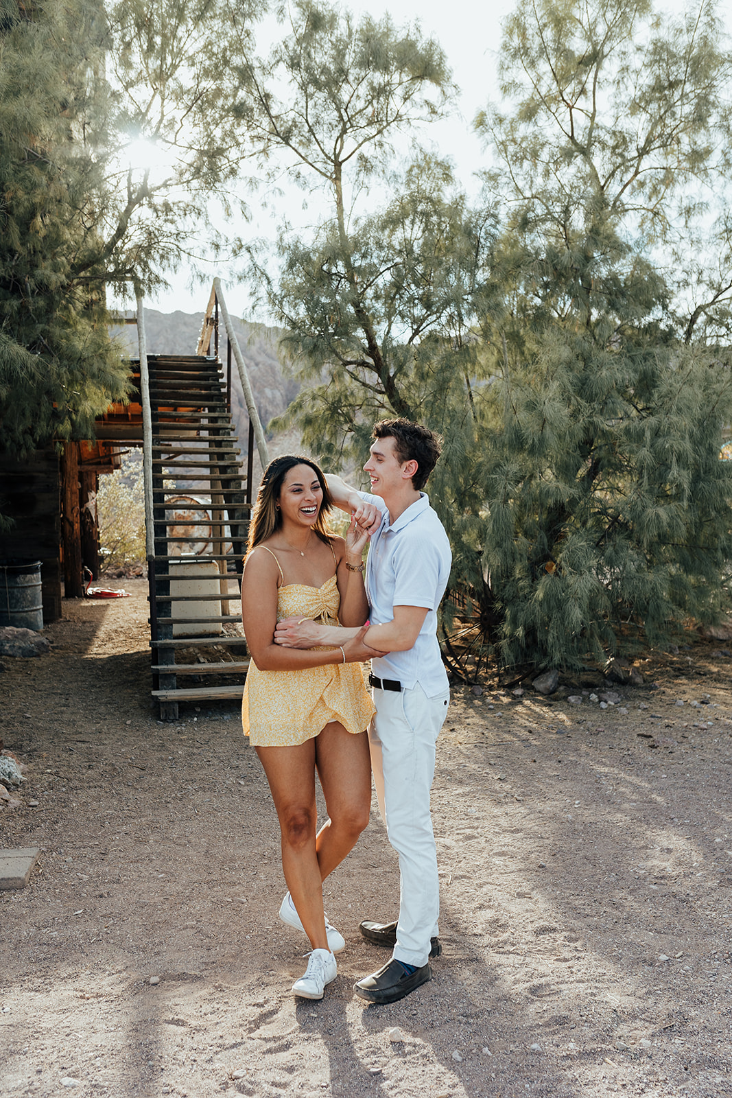 A couple embracing and smiling at Nelson's Ghost Town, Nevada, with desert trees, a wooden staircase, and mountains in the background.