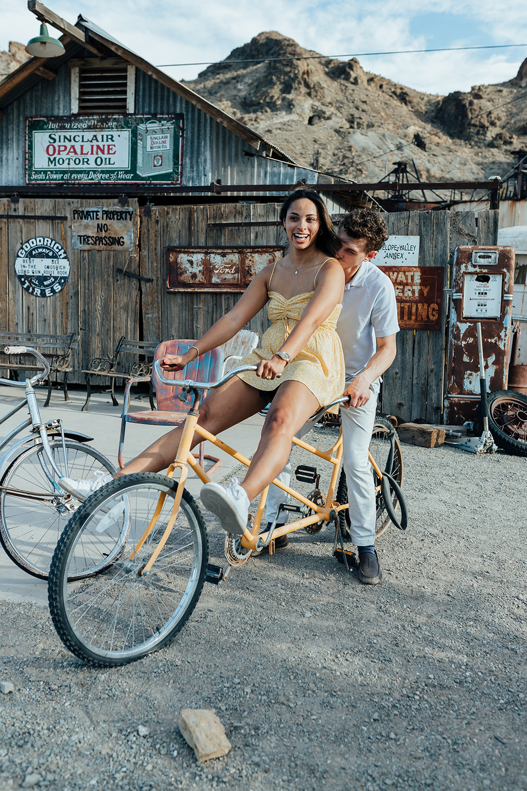 A couple riding a tandem bike at Nelson's Ghost Town, Nevada, with vintage signs, rusted props, and desert mountains in the background.