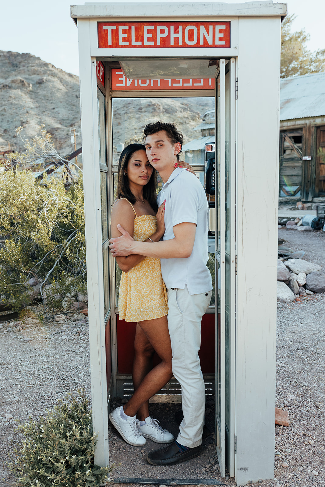 A couple embracing inside a vintage telephone booth at Nelson's Ghost Town, Nevada, with desert mountains and rustic structures in the background.