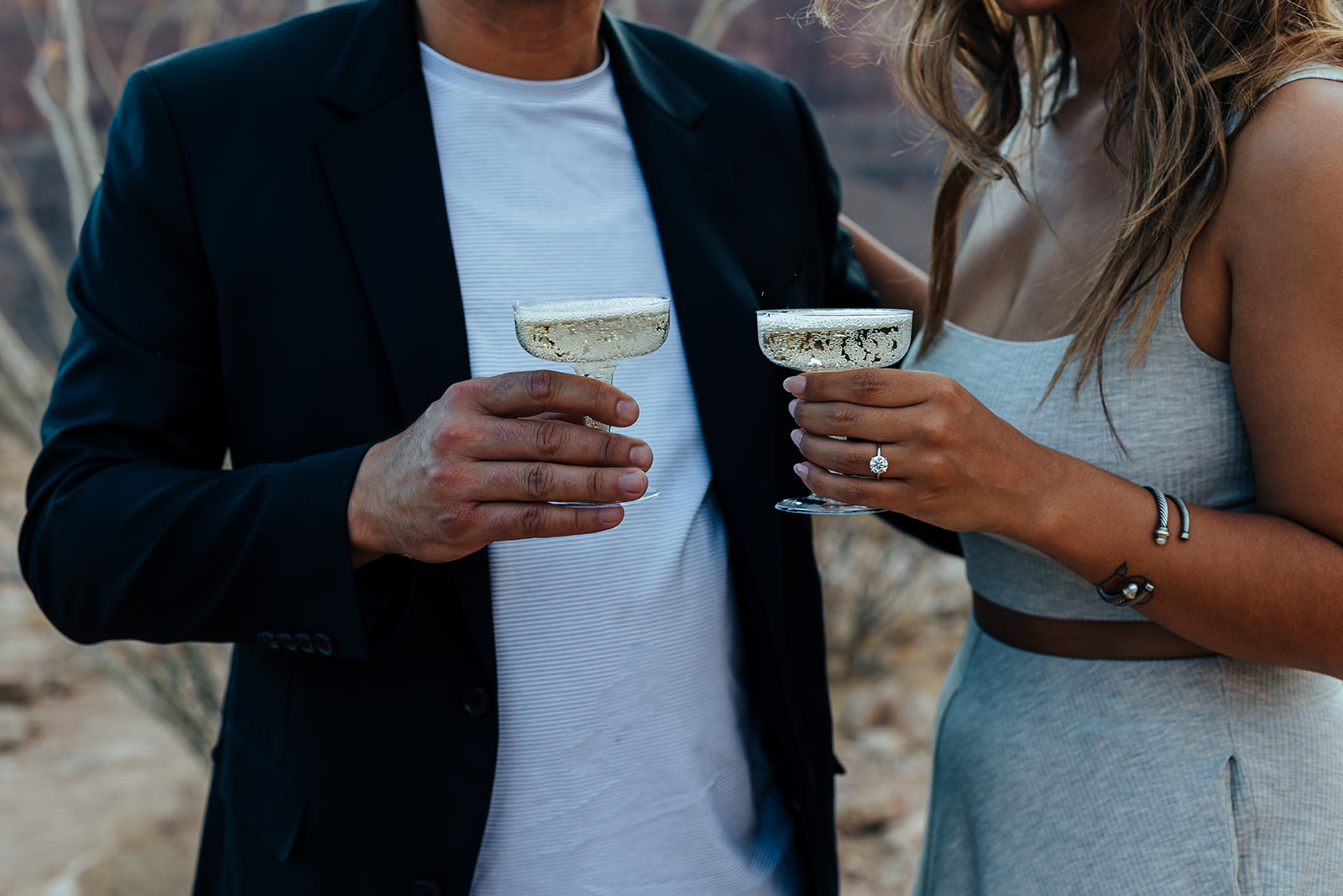 A couple toasts with champagne glasses in the Grand Canyon's rugged landscape during a helicopter proposal. The woman, with long hair and a ring, wears a white top and skirt, while the man in a black suit stands close, both adorned with bracelets, against a backdrop of rocky terrain.