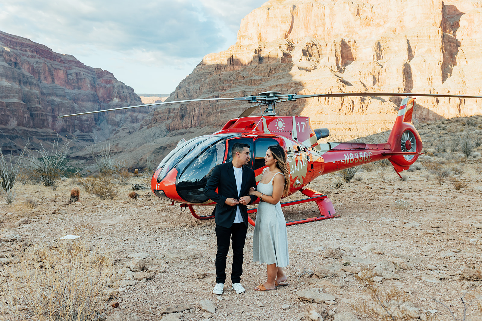 A couple stands together in front of a red helicopter in the Grand Canyon's rocky terrain during a proposal. The woman, with long hair, wears a white dress and sandals, while the man in a black suit and white sneakers adjusts his jacket, with the canyon's layered cliffs in the background.
