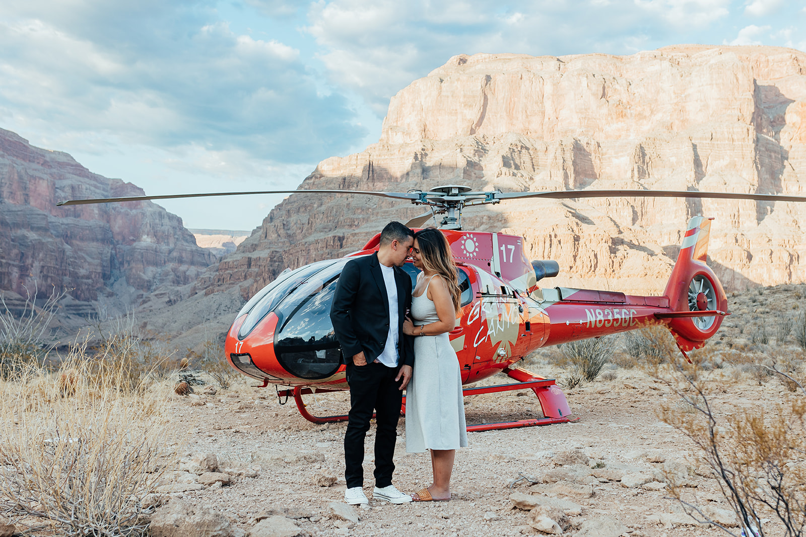A couple shares an intimate moment, touching foreheads, in front of a red helicopter in the Grand Canyon's rugged landscape during a proposal. The woman, with long hair, wears a white dress and sandals, while the man in a black suit and white sneakers stands close, with towering cliffs and a partly cloudy sky behind them.