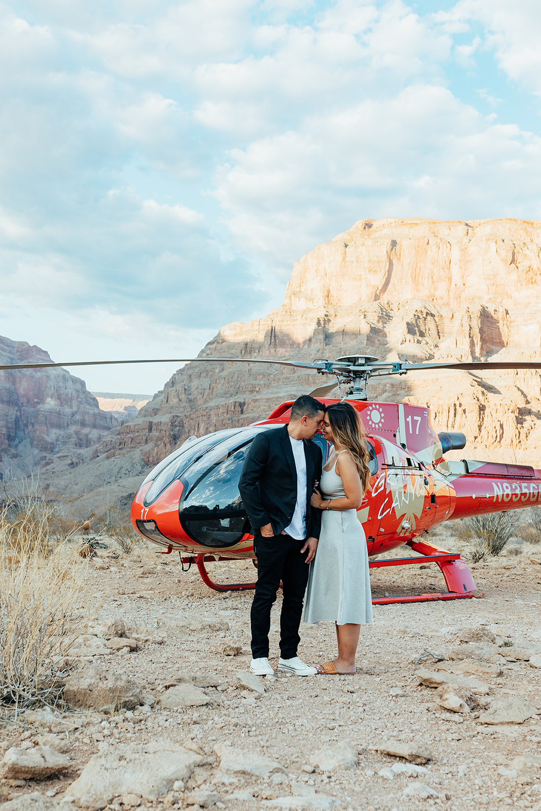 A couple embraces in front of a red helicopter in the Grand Canyon's rocky terrain during a proposal, with towering cliffs and a partly cloudy sky in the background. The woman, with long hair, wears a white dress and sandals, while the man in a black suit and white sneakers holds her close.