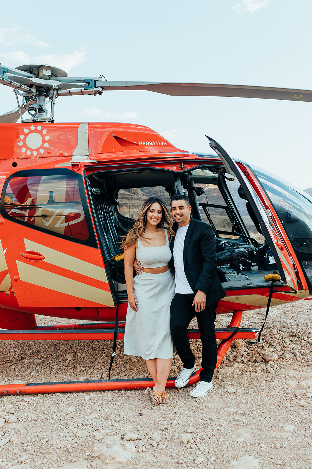 A couple stands happily by a red helicopter with an open door in the Grand Canyon's rocky terrain during a proposal. The woman, with long hair, wears a white dress and sandals, while the man in a black suit and white sneakers embraces her, with distant cliffs and a partly cloudy sky in the background.