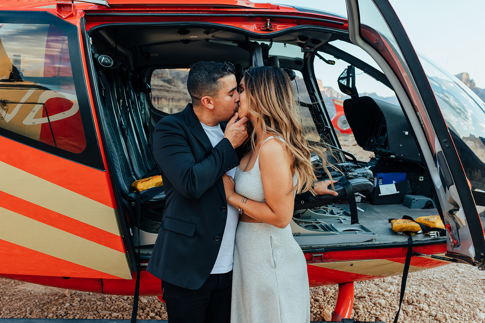 A couple shares a kiss in front of a red helicopter with an open door in the Grand Canyon's rocky terrain during a proposal. The woman, with long hair and a bracelet, wears a white dress, while the man in a black suit embraces her, with distant cliffs visible in the background.