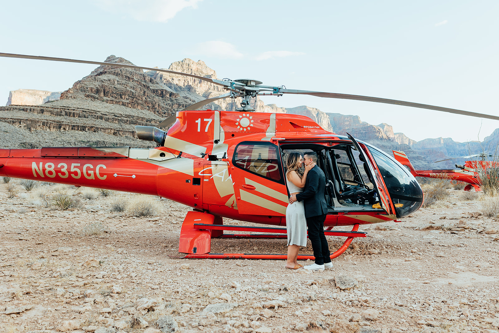 A couple shares a kiss inside a red helicopter in the Grand Canyon's rocky terrain during a proposal, with the door open and towering cliffs in the background. The woman, with long hair, wears a white dress and sandals, while the man in a black suit and white sneakers embraces her.