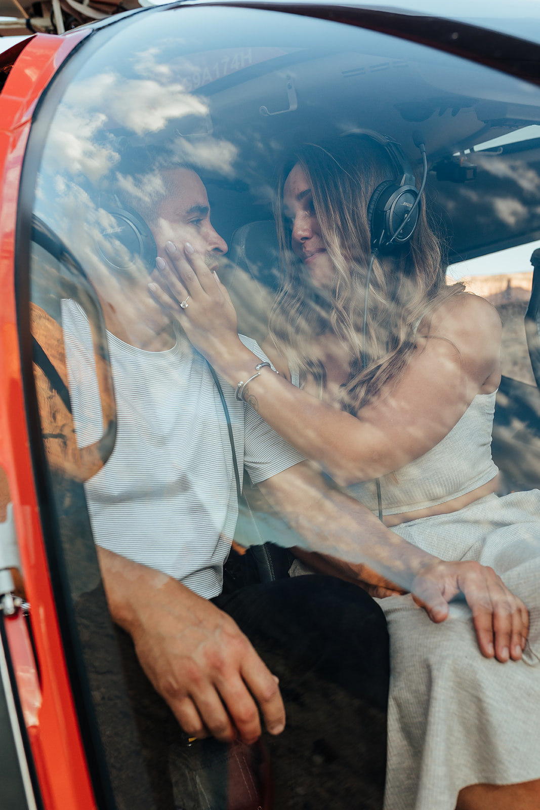 A couple shares a tender moment inside a red helicopter in the Grand Canyon's terrain during a proposal, with the woman touching the man's face while wearing headsets. She has long hair, a bracelet, and a white dress, while he wears a white shirt and black suit, with cliffs visible through the window.