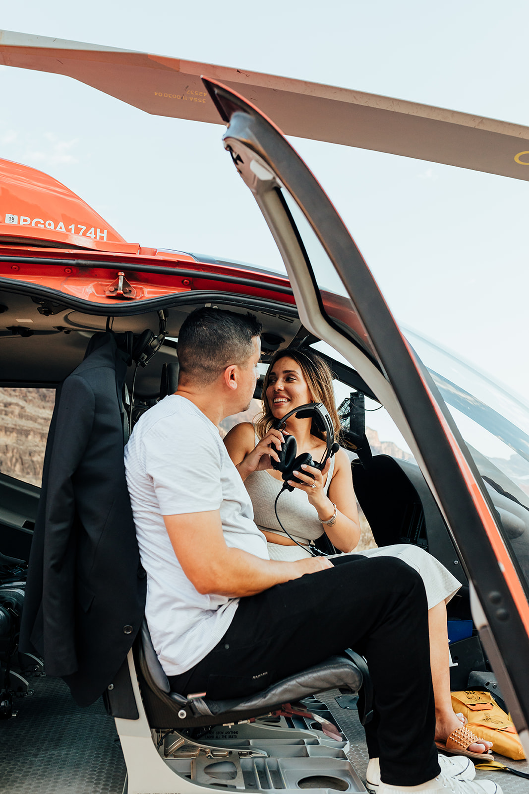 A couple sits inside a red helicopter in the Grand Canyon's terrain during a proposal, smiling at each other with headsets on. The woman, with long hair and a bracelet, wears a white dress, while the man in a white shirt and black pants looks at her, with a jacket hanging behind them.