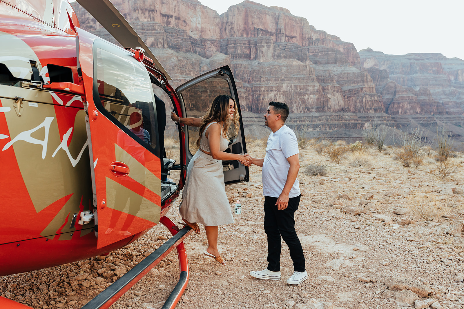 A couple shakes hands by a red helicopter in the Grand Canyon's rocky terrain during a proposal, with the door open and towering cliffs in the background. The woman, with long hair, wears a white dress and sandals, while the man in a white shirt and black pants stands beside her.