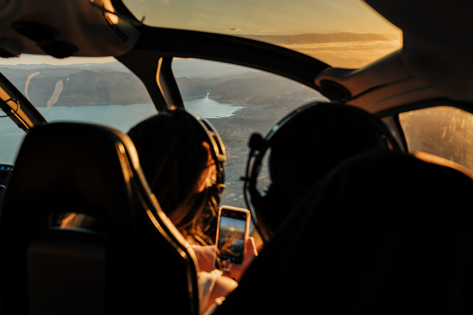 A couple in a helicopter over the Grand Canyon during sunset, with one person taking a photo on their phone. The stunning landscape features rugged cliffs and a serene lake, capturing the moment of a romantic proposal.