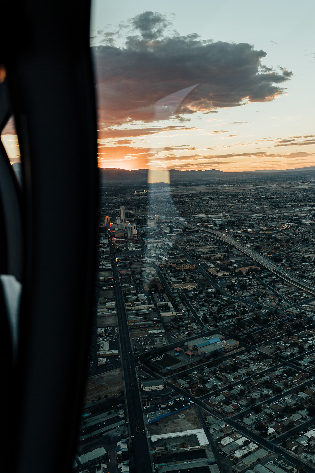 A breathtaking sunset view of a cityscape from a helicopter, with the sun casting an orange glow over buildings and highways. The distant mountains hint at the Grand Canyon's proximity, setting the scene for a romantic helicopter proposal.