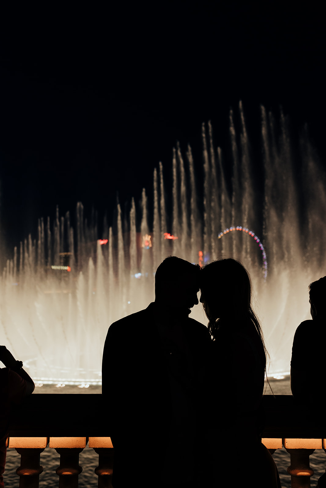 A couple's silhouette against the illuminated Bellagio Fountains at night, sharing a tender moment with foreheads touching. The colorful Ferris wheel in the background adds to the romantic ambiance in Las Vegas, following their Grand Canyon helicopter proposal.