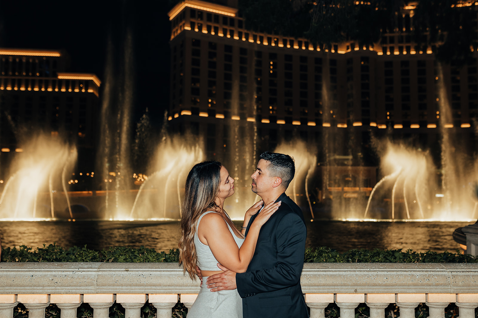 A couple embraces in front of the glowing Bellagio Fountains at night, with the man in a suit and the woman in a white outfit. The illuminated Bellagio hotel in the background enhances the romantic setting in Las Vegas, celebrating their Grand Canyon helicopter proposal.