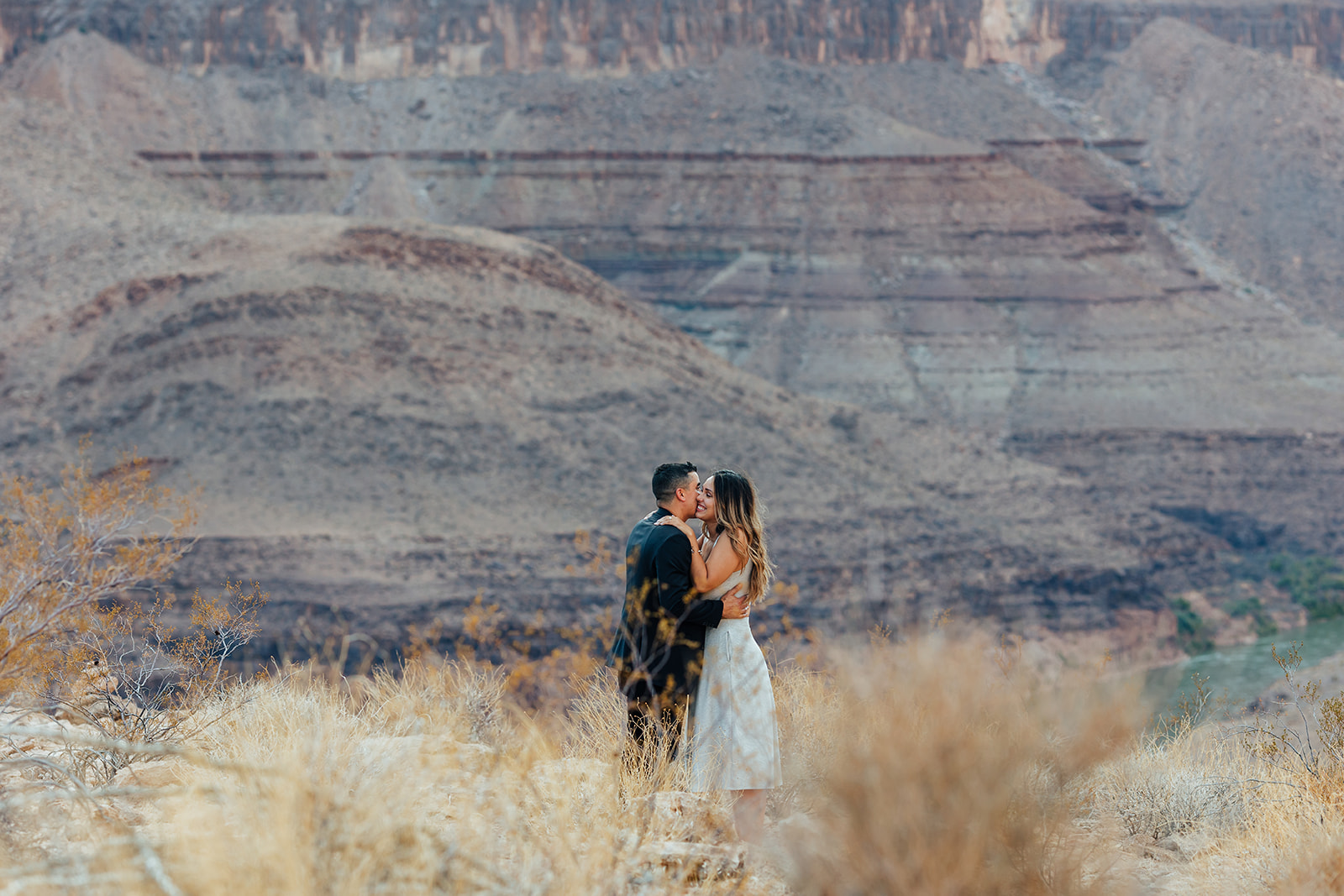 A couple shares a kiss on a Grand Canyon ledge after a helicopter proposal, surrounded by dry grass with layered canyon cliffs and a river in the background.