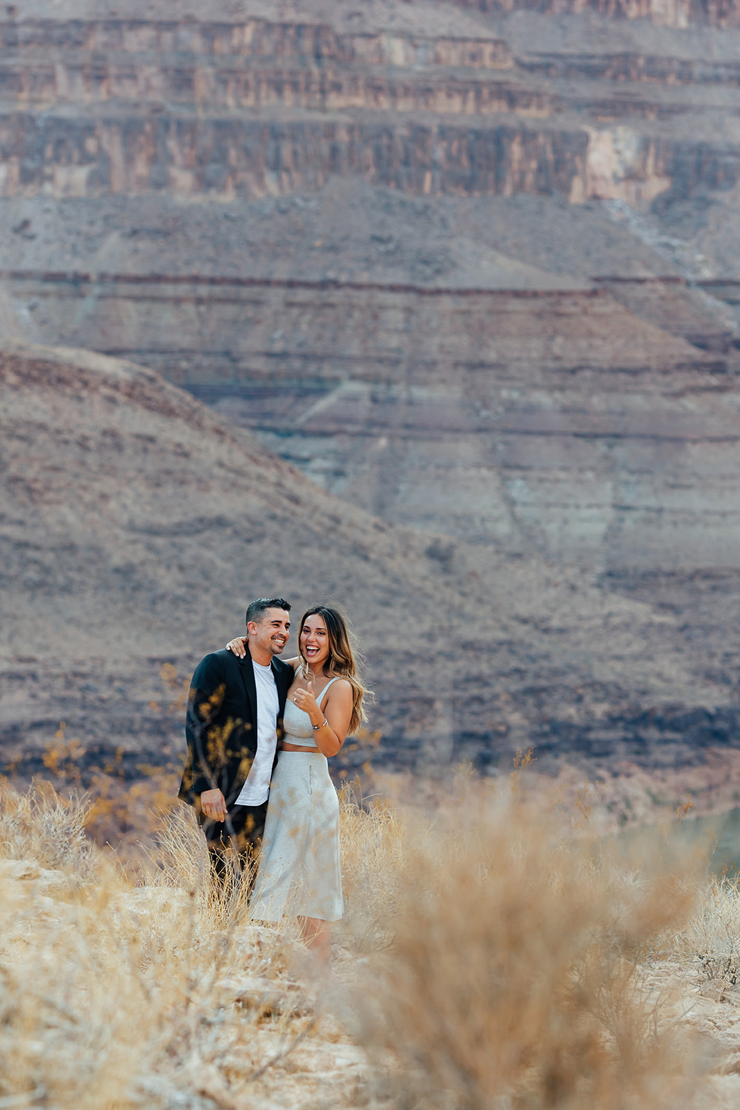 A couple shares a joyful moment on a Grand Canyon ledge after a helicopter proposal, surrounded by dry grass with layered canyon cliffs in the background.