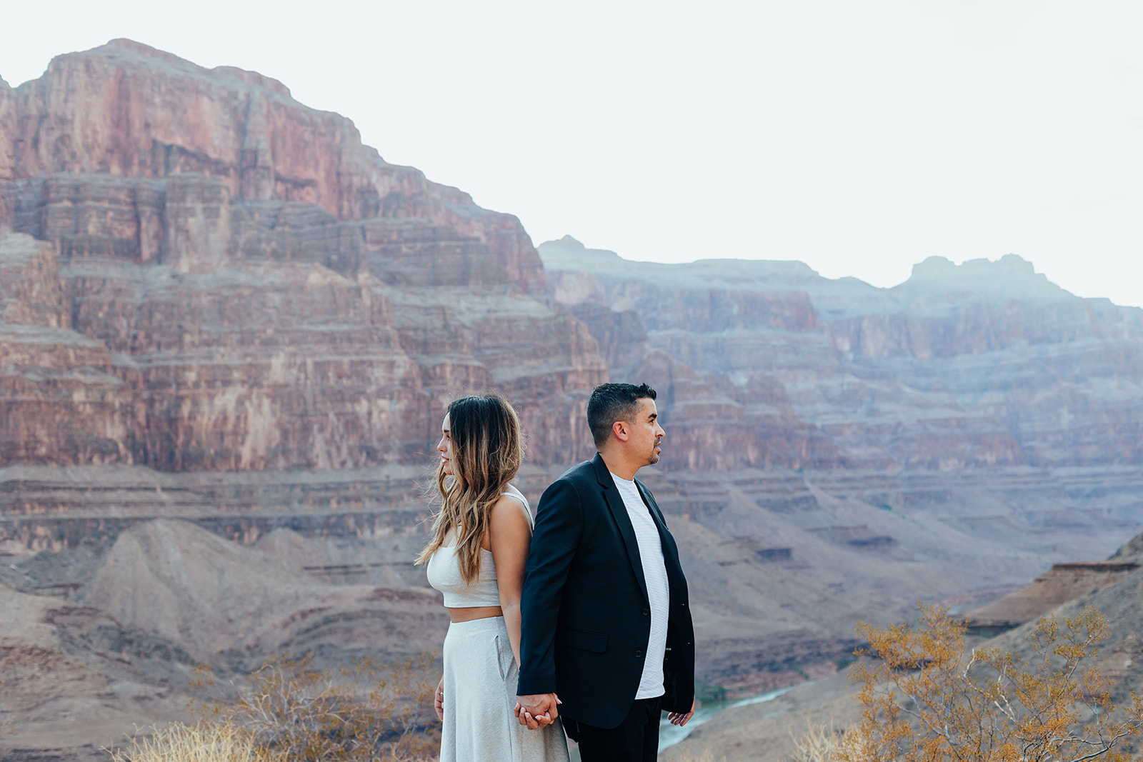 A couple stands hand-in-hand on a Grand Canyon ledge after a helicopter proposal, with layered canyon cliffs and a river in the background.