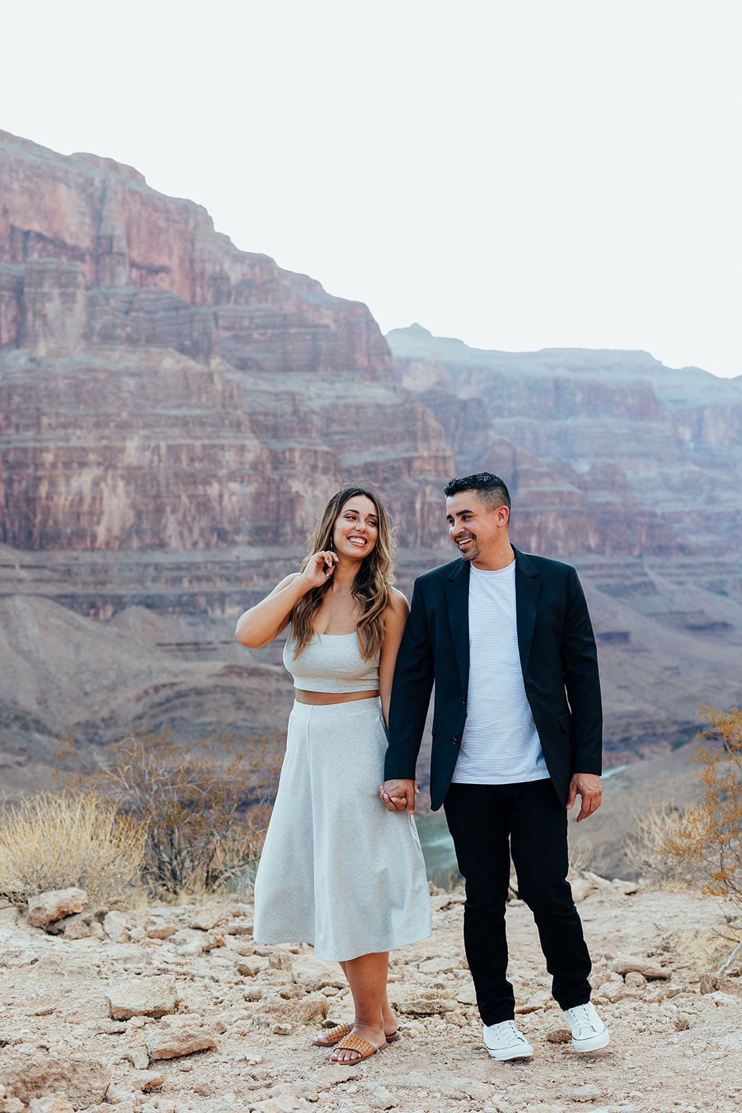 A couple smiles and holds hands on a Grand Canyon ledge after a helicopter proposal, with layered canyon cliffs and a river in the background.