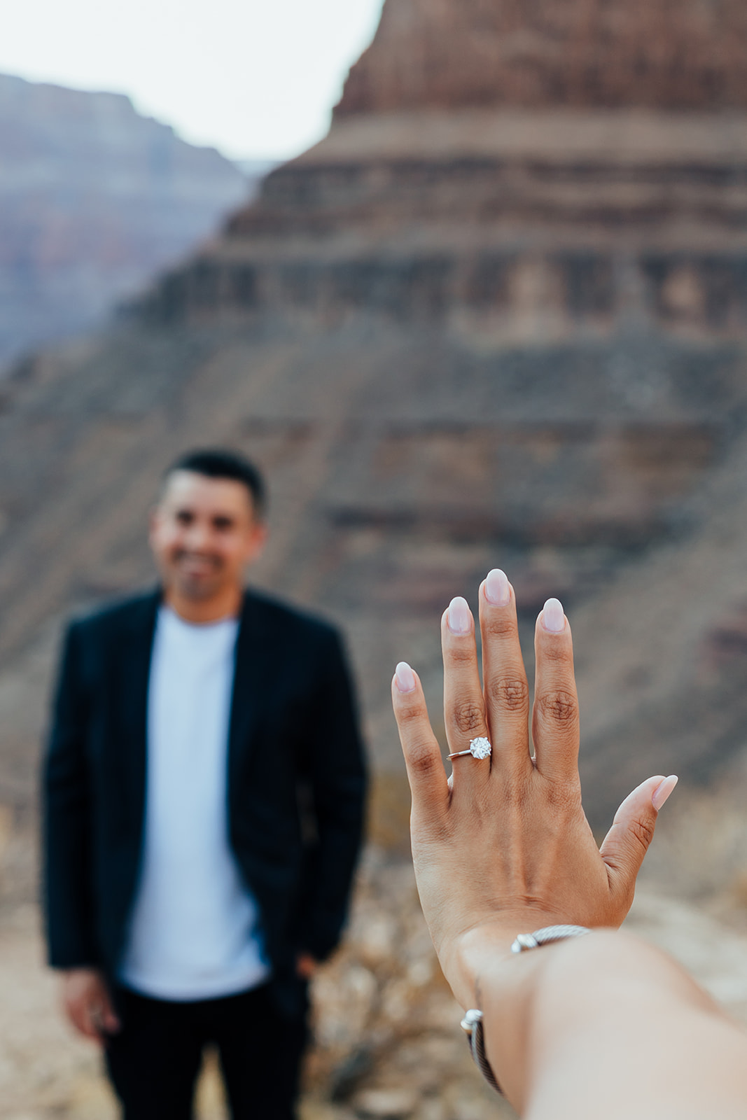 A hand with a new engagement ring is shown in the foreground, with a man in a suit standing on a Grand Canyon ledge and layered canyon cliffs in the background after a helicopter proposal.