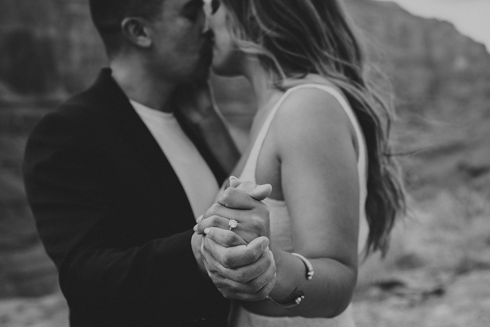 A black-and-white close-up of a couple holding hands, with a new engagement ring visible, against a blurred Grand Canyon backdrop after a helicopter proposal.