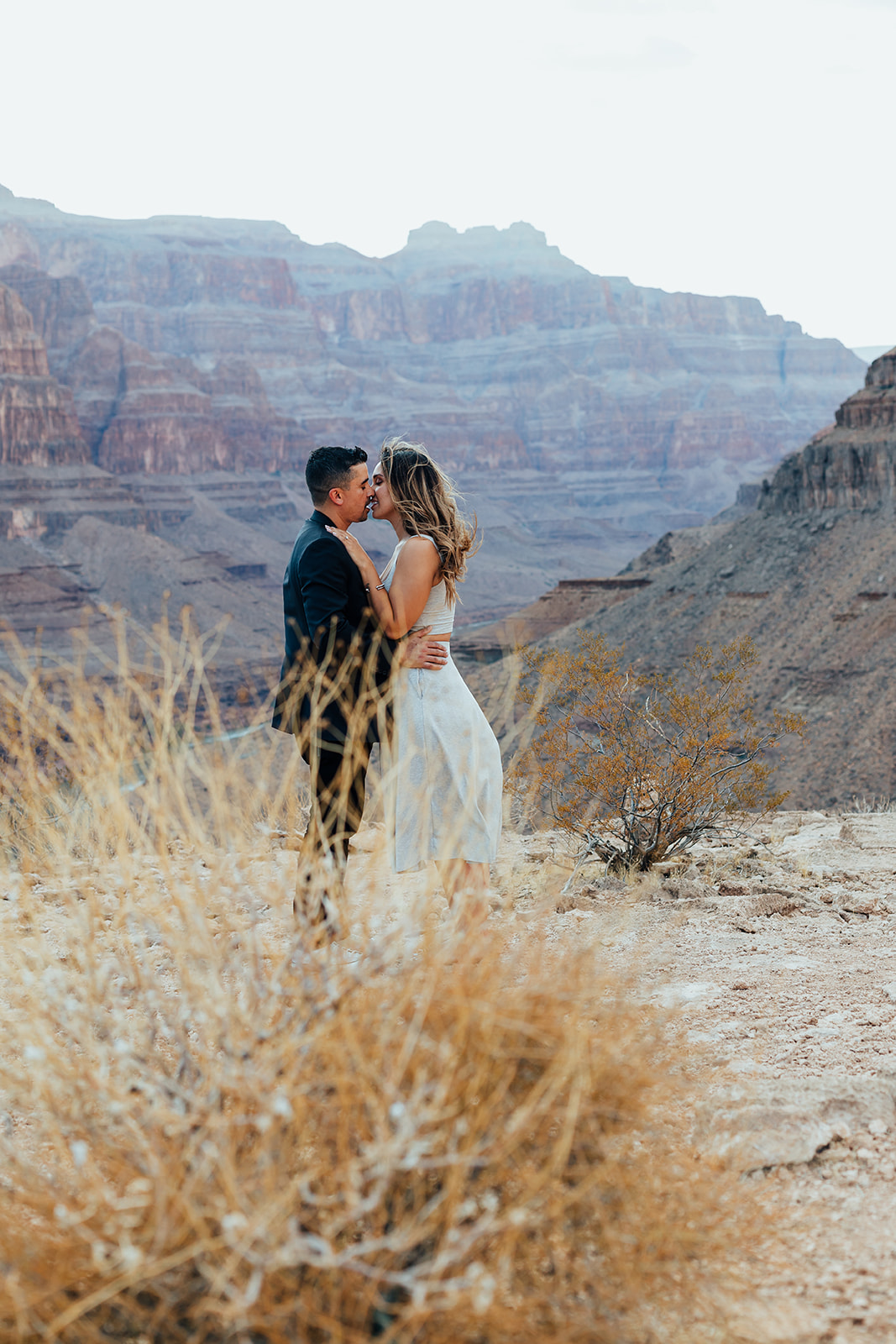 A couple shares a romantic kiss on a Grand Canyon ledge after a helicopter proposal, with layered canyon cliffs in the background and dry grass in the foreground.