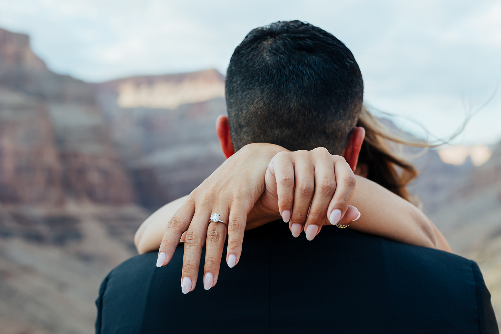 A couple embraces with the woman's arms around the man's shoulders, her new engagement ring visible, against a Grand Canyon backdrop after a helicopter proposal.
