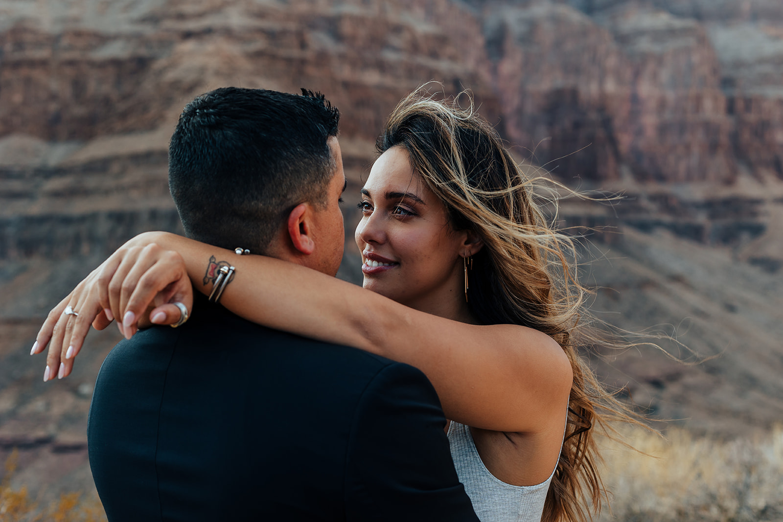 A couple embraces in front of the Grand Canyon's rugged cliffs during a helicopter proposal. The woman, with long, wind-swept hair, wears a white top and earrings, while the man in a black suit holds her close, showcasing a ring on her finger and bracelets on her wrist.