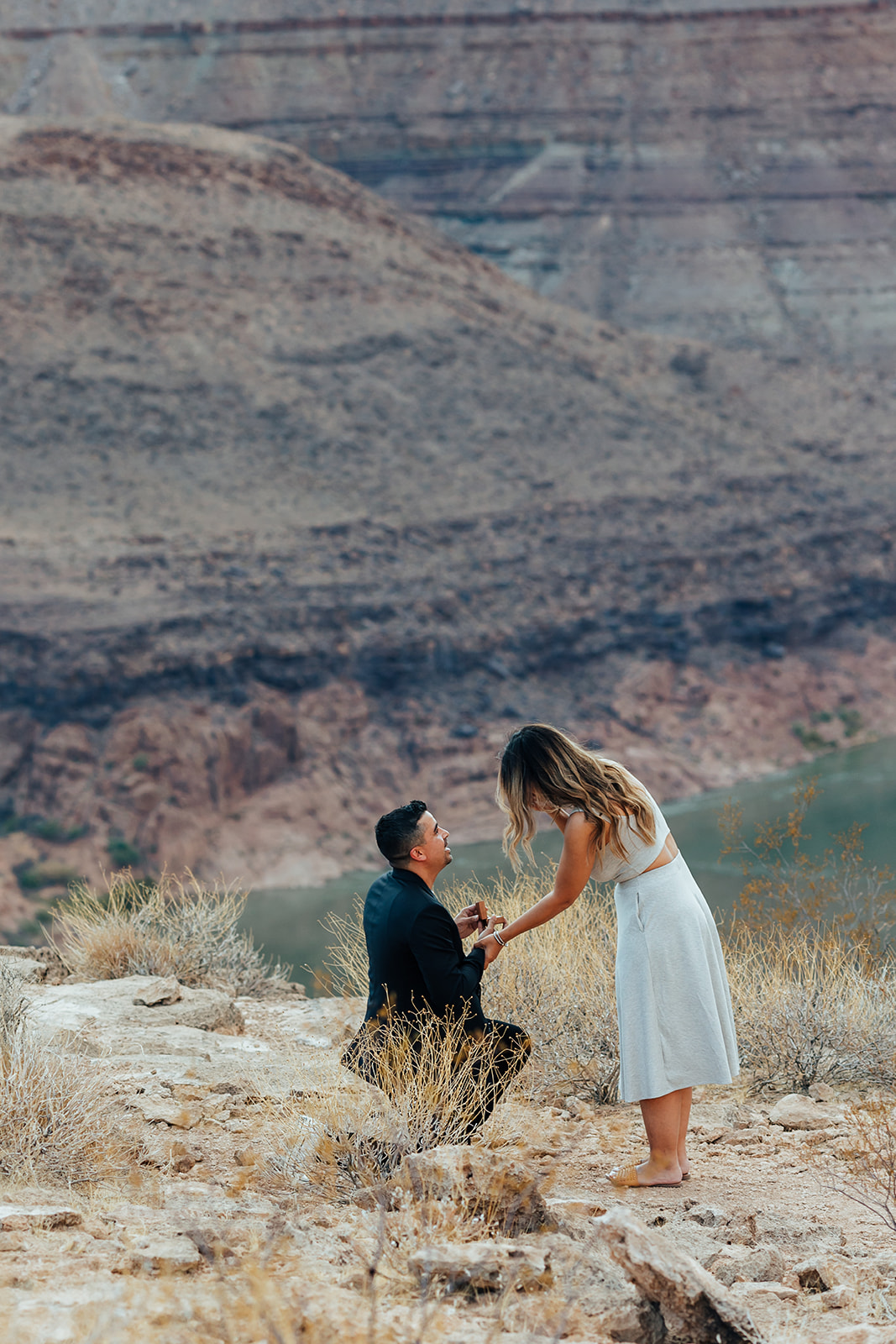 A man kneels to propose to a woman in a white outfit on a rocky Grand Canyon ledge, with layered canyon cliffs and a river in the background, captured after a helicopter arrival.