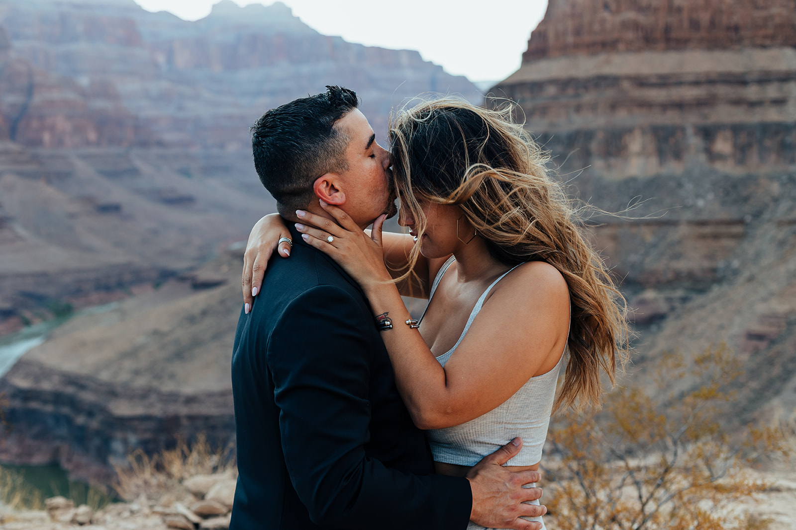 A couple shares a tender kiss with the Grand Canyon's dramatic cliffs and river in the background during a helicopter proposal. The woman, with long, flowing hair, wears a white top and a ring, while the man in a black suit embraces her, their hands adorned with bracelets and rings.