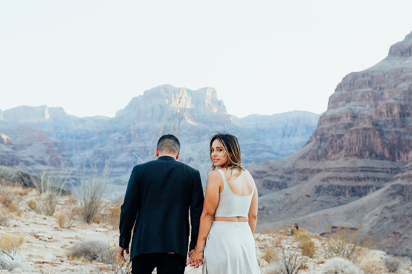 A couple holds hands, facing away from the camera, with the Grand Canyon's majestic cliffs in the background during a helicopter proposal. The woman, with long hair, wears a white top and skirt, while the man in a black suit walks beside her, both adorned with bracelets and rings.