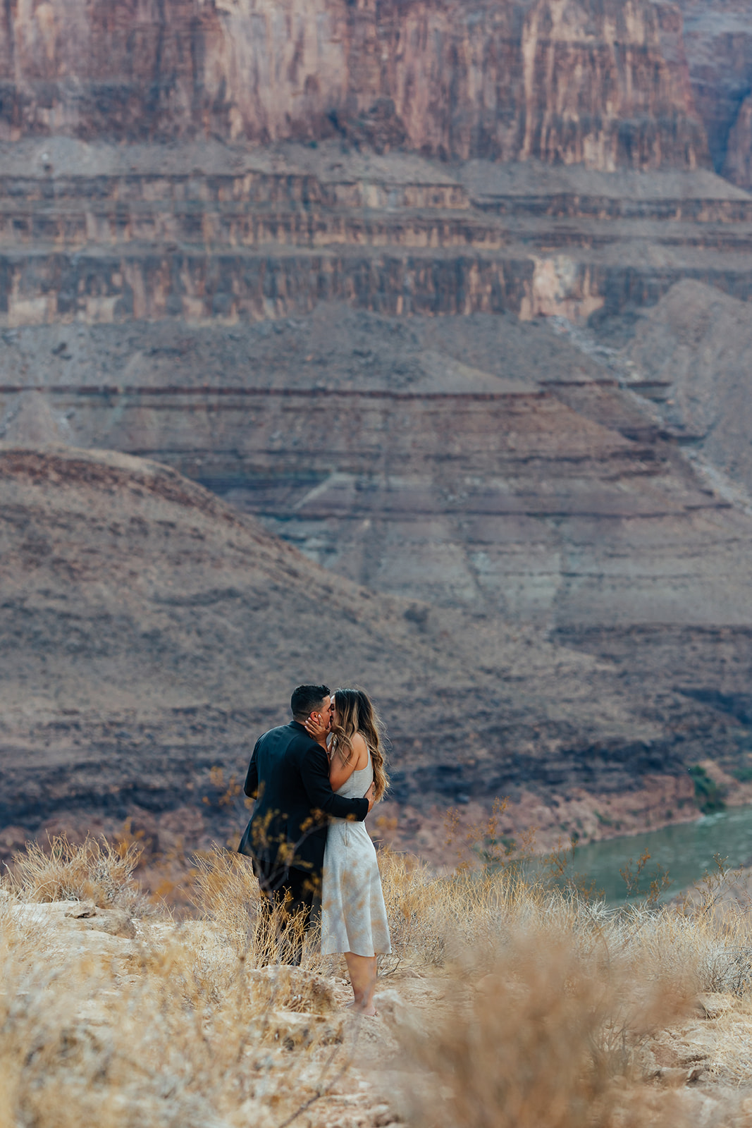 A couple embraces on a Grand Canyon ledge after a helicopter proposal, with layered canyon cliffs and a river in the background.